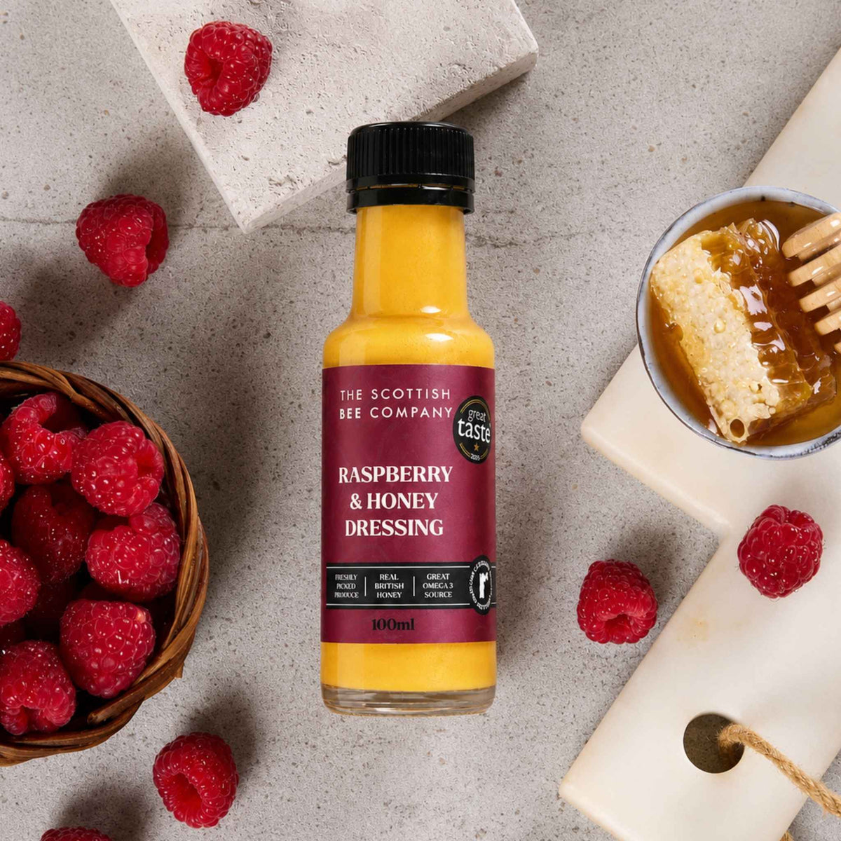 A bottle of The Scottish Bee Company Raspberry & Honey Dressing is styled in a flat-lay arrangement with fresh raspberries, a bowl of honey, and stone tiles on a textured gray background.