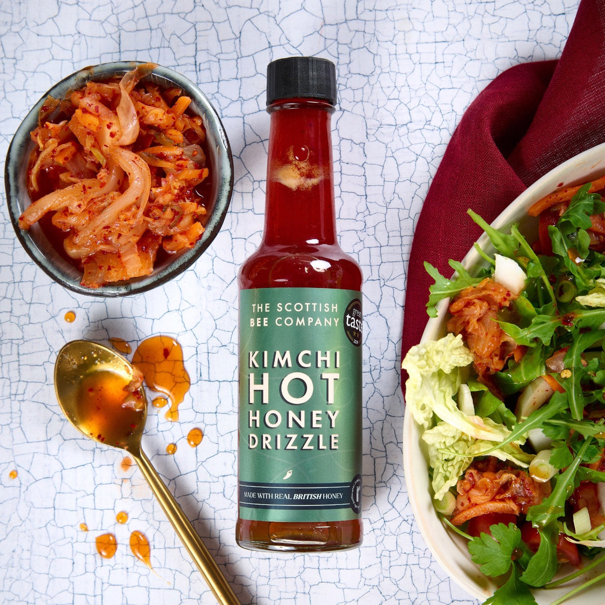 Bottle of Kimchi Hot Honey Drizzle with a bowl of kimchi and salad on a textured surface